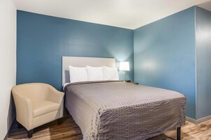 A hotel room with a bed, a chair, a nightstand with a lamp, and a clock.