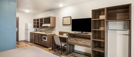 A compact hotel room with a kitchenette, a bed, a desk with a chair, a TV, and built-in shelving.