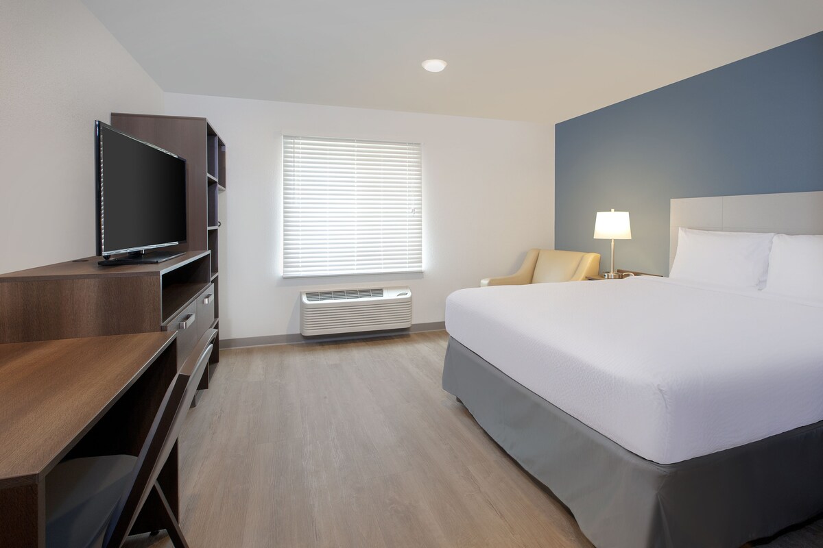 A hotel room with a large bed, a TV on a wooden dresser, a window with blinds, a bedside lamp, and a wall-mounted heater.