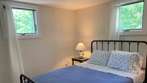 Un dormitorio con cama, mesita de noche con lámpara y dos ventanas con cortinas.