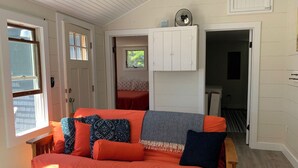 Una habitación acogedora con piso de madera, un sofá con cojines naranjas y estampados, un armario blanco y una ventana con persianas.