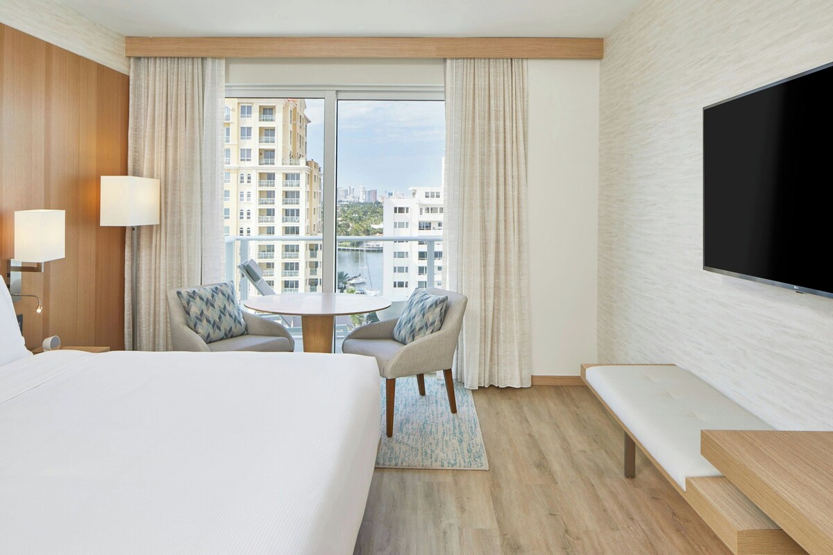 Une chambre d’hôtel avec un grand lit, une télévision, une table à manger avec des chaises et une vue sur les bâtiments par la fenêtre.