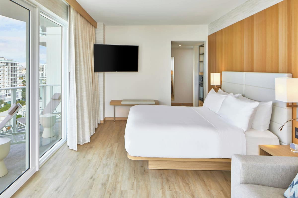 Habitación de hotel con una cama grande, televisor montado en la pared, balcón con asientos y cabecera de madera.