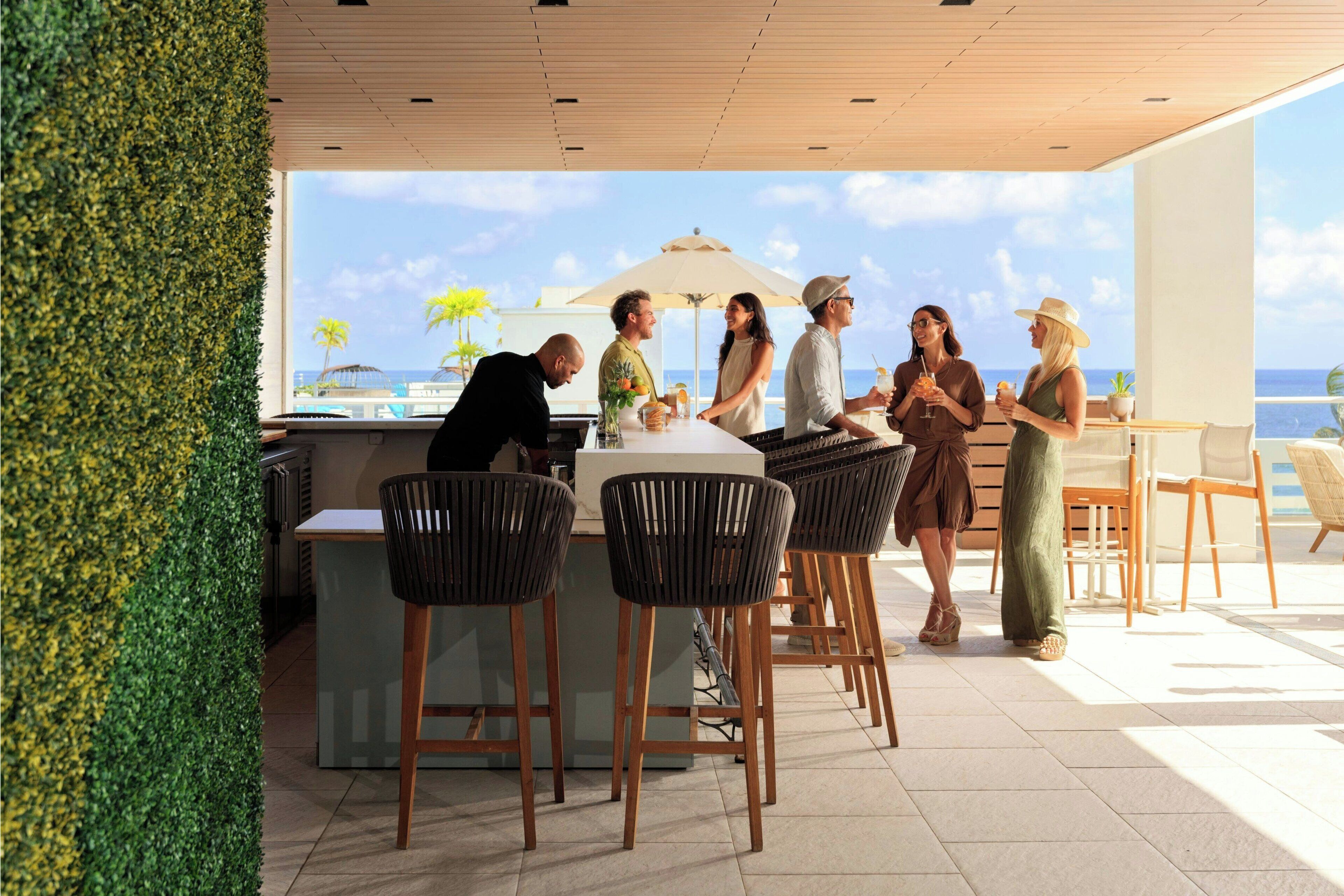 A modern outdoor dining area with a bar, wooden chairs, and a view of the ocean.