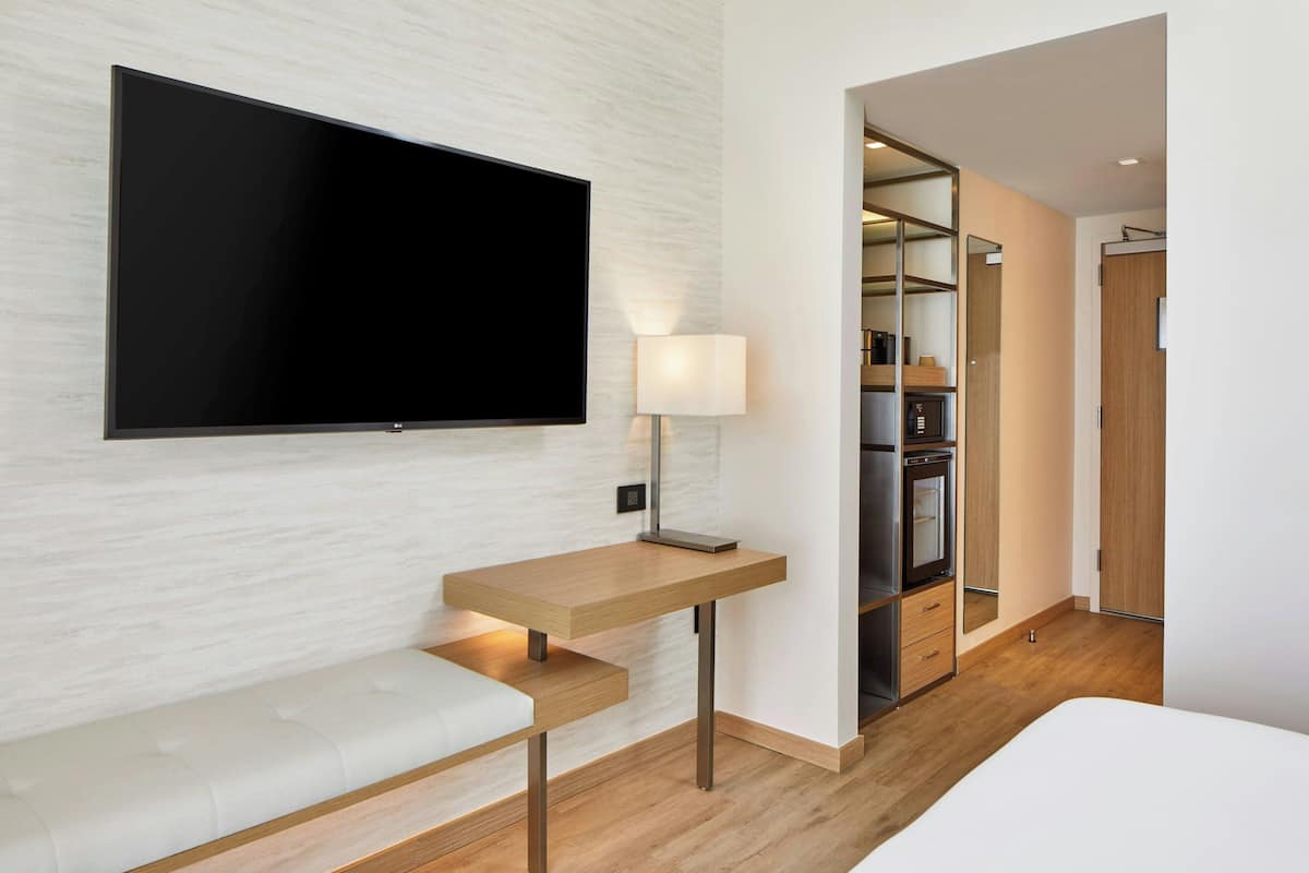 Una habitación de hotel moderna con una televisión de pantalla plana montada en la pared, una mesita de noche con lámpara, un banco y un vestidor con estantes y espejo.
