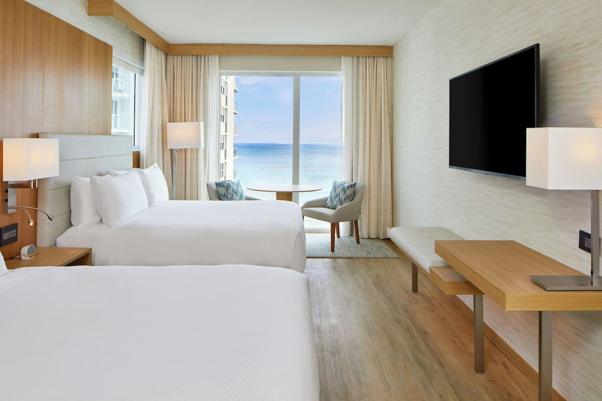 Habitación de hotel con dos camas, televisión, escritorio y vista al mar.