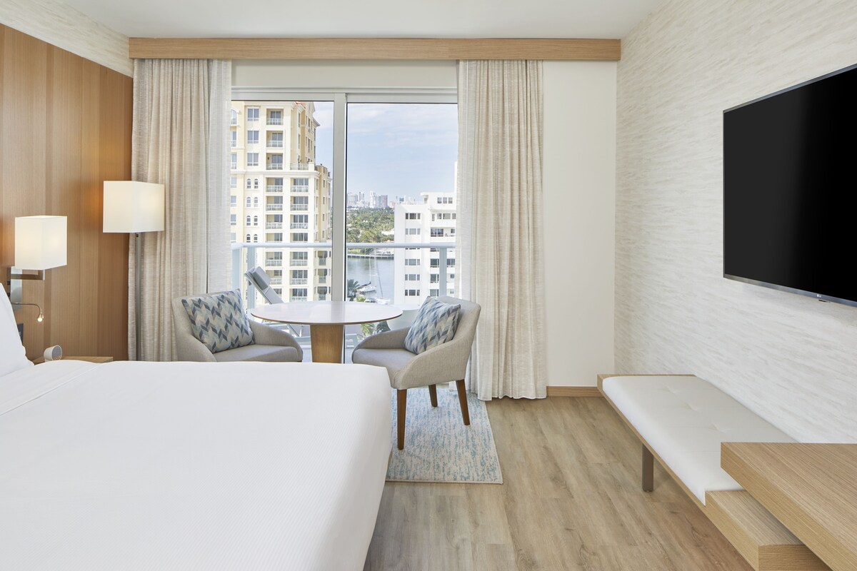 Ein Hotelzimmer mit einem großen Bett, einem runden Tisch mit zwei Stühlen, einem an der Wand befestigten Fernseher und Blick auf die Stadt durch das Fenster.