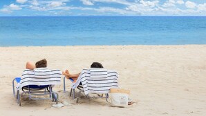 Zwei Personen sitzen in Strandkörben mit gestreiften Handtüchern, einem Strandbeutel und Blick auf das Meer.