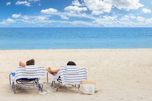 Deux personnes sont assises sur des chaises de plage, entourées de serviettes à rayures et d’un sac de plage, et elles ont une vue dégagée sur l’océan.