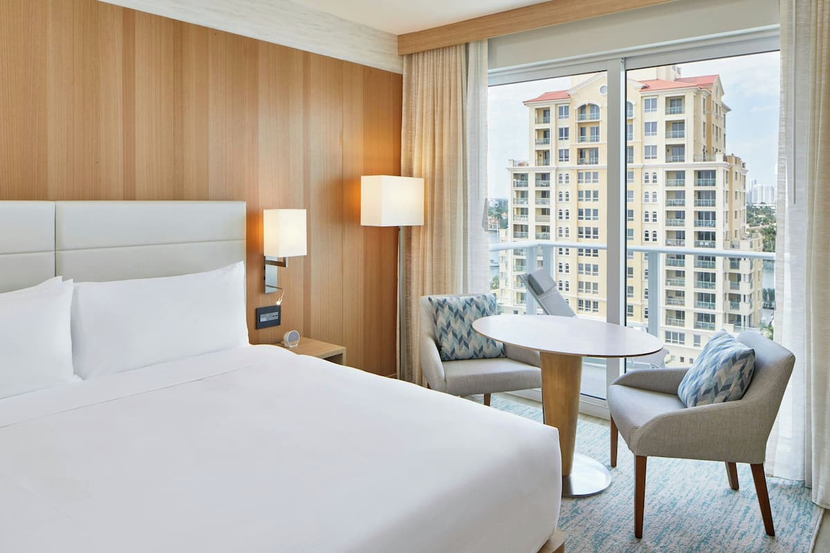 Habitación de hotel con una cama grande, una mesita pequeña, una silla y vistas a un rascacielos.