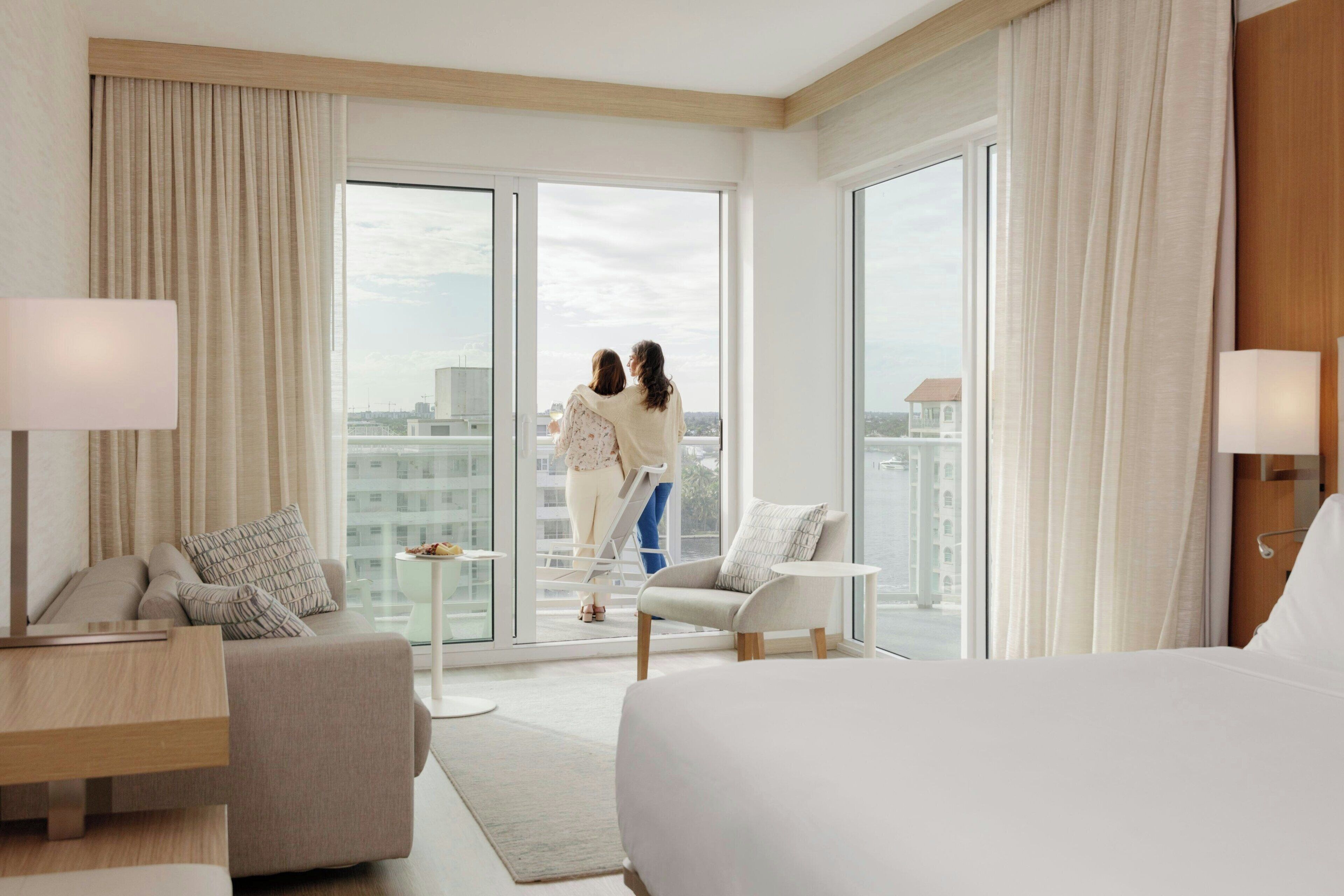 A modern hotel room with a large bed, a sofa, a small table, and a view of the city through large windows.
