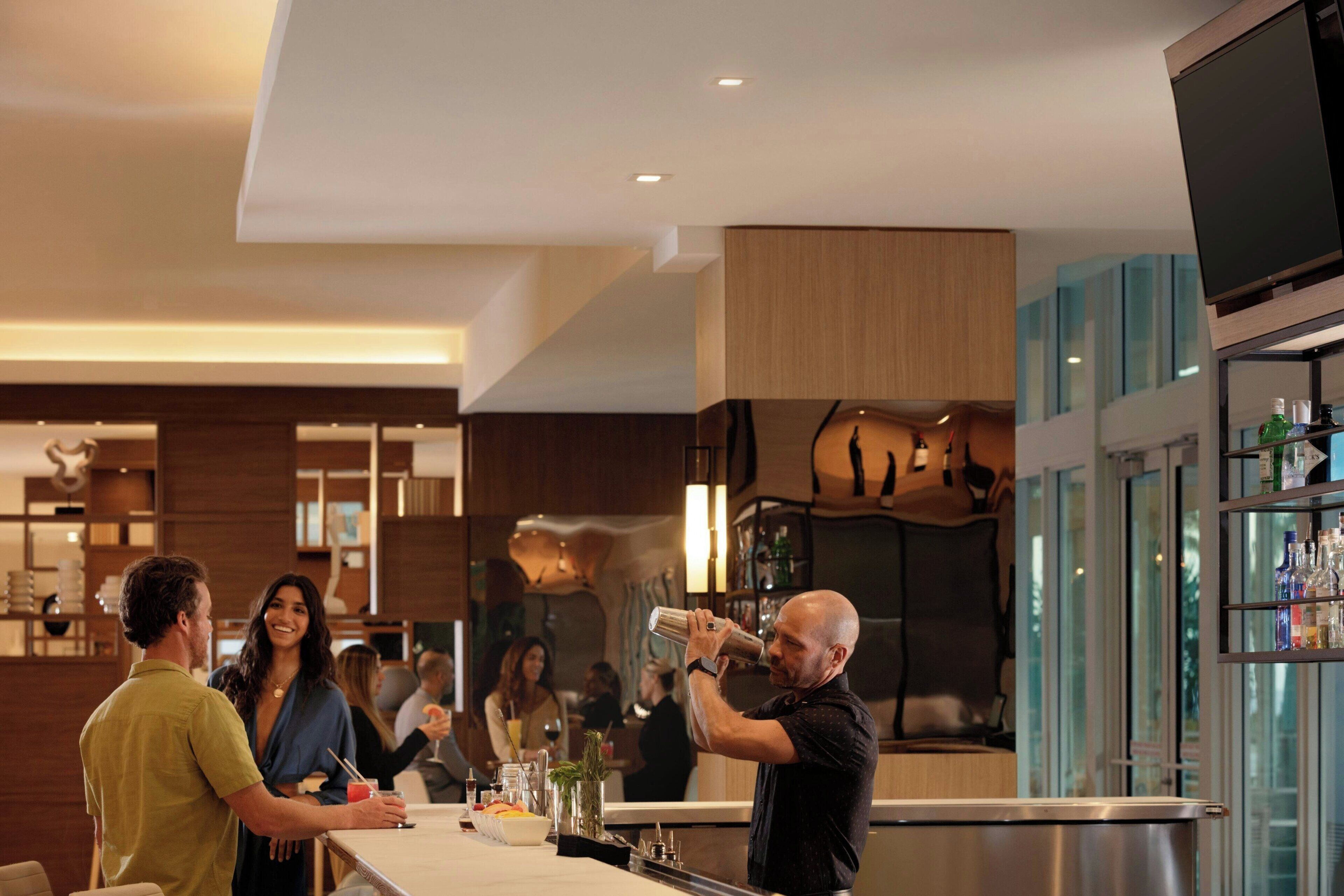 A modern bar area with a bartender preparing drinks, a counter with various items, and a glass display case with bottles.