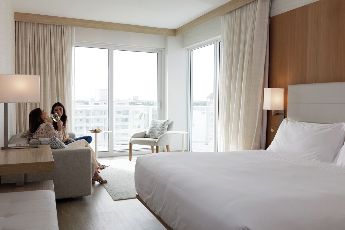 Ein Hotelzimmer mit einem großen Bett, einem Sofa, einem Sessel und einem kleinen Tisch. Zwei Fenster mit Vorhängen bieten einen Blick auf umliegende Gebäude.