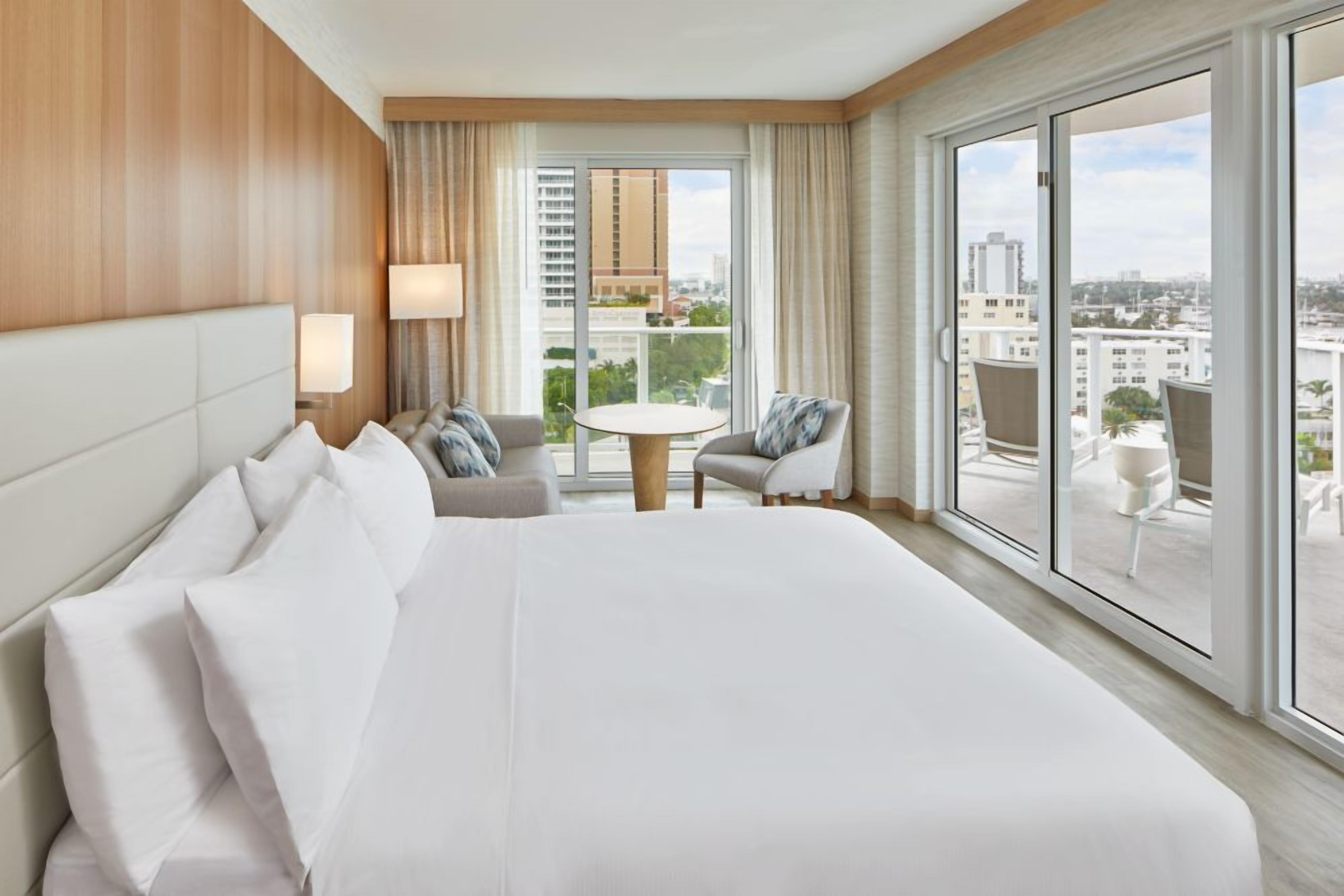 Une chambre d’hôtel avec un grand lit, une table ronde, deux chaises et une vue sur la ville grâce à de grandes fenêtres.