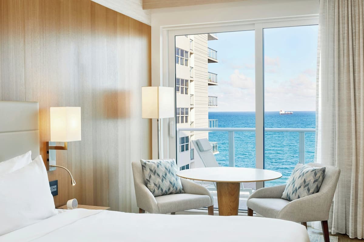 Habitación de hotel con cama, dos sillones, una mesa redonda y un amplio ventanal con vistas al océano y un crucero.