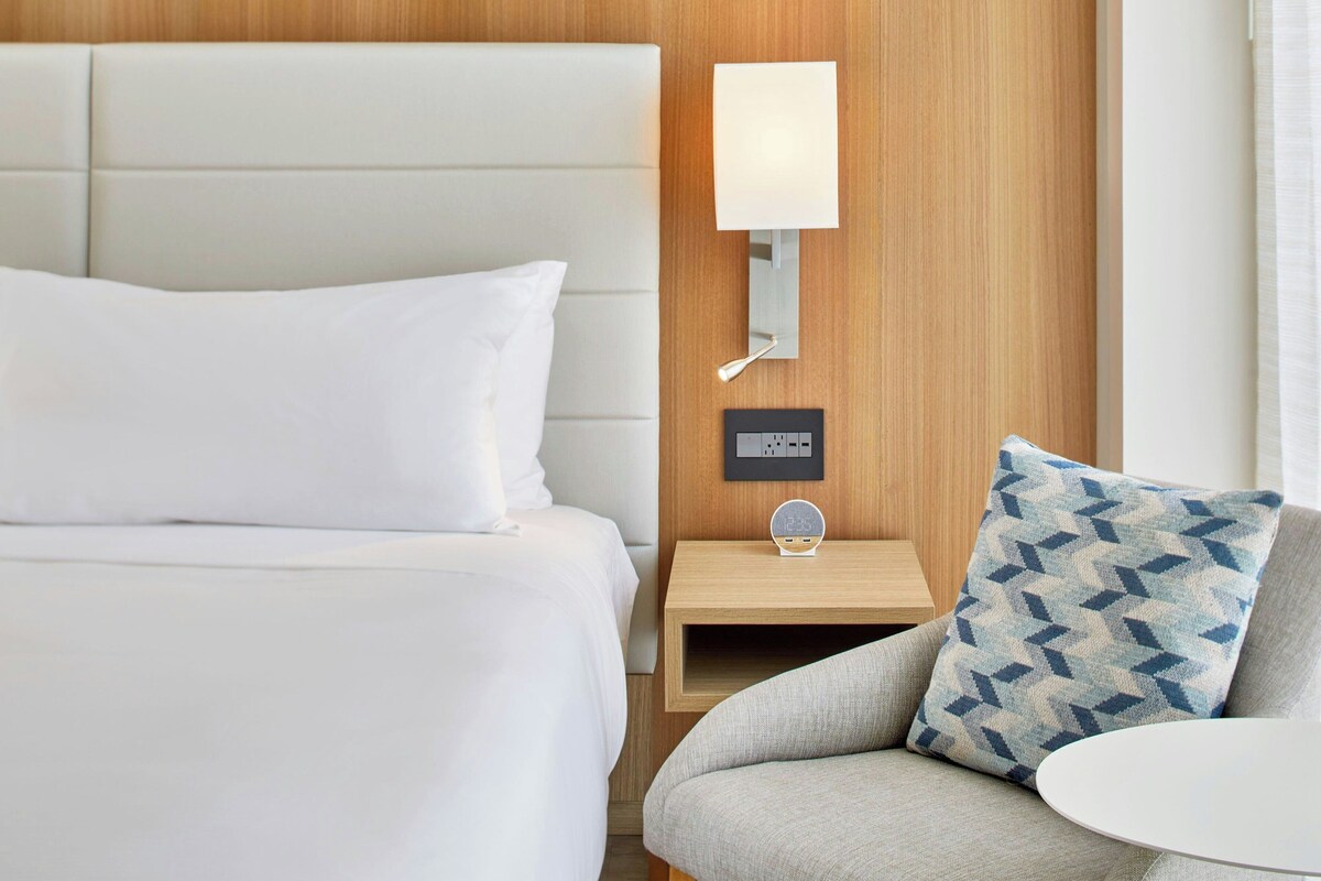 Ein Hotelzimmer mit Bett, Nachttisch mit Lampe, digitaler Uhr und Kissen mit Muster.