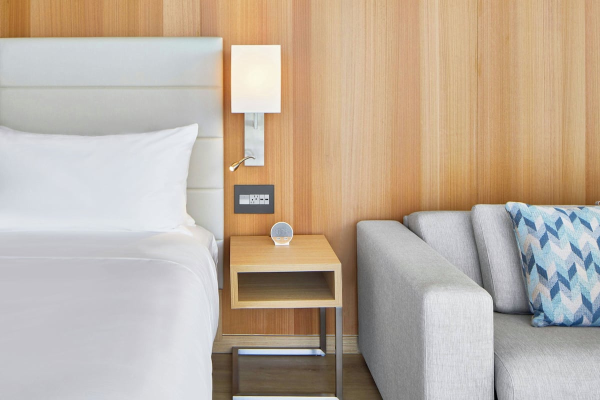 Una habitación de hotel con una cama, una mesita de noche con lámpara, un estante pequeño y un sofá.