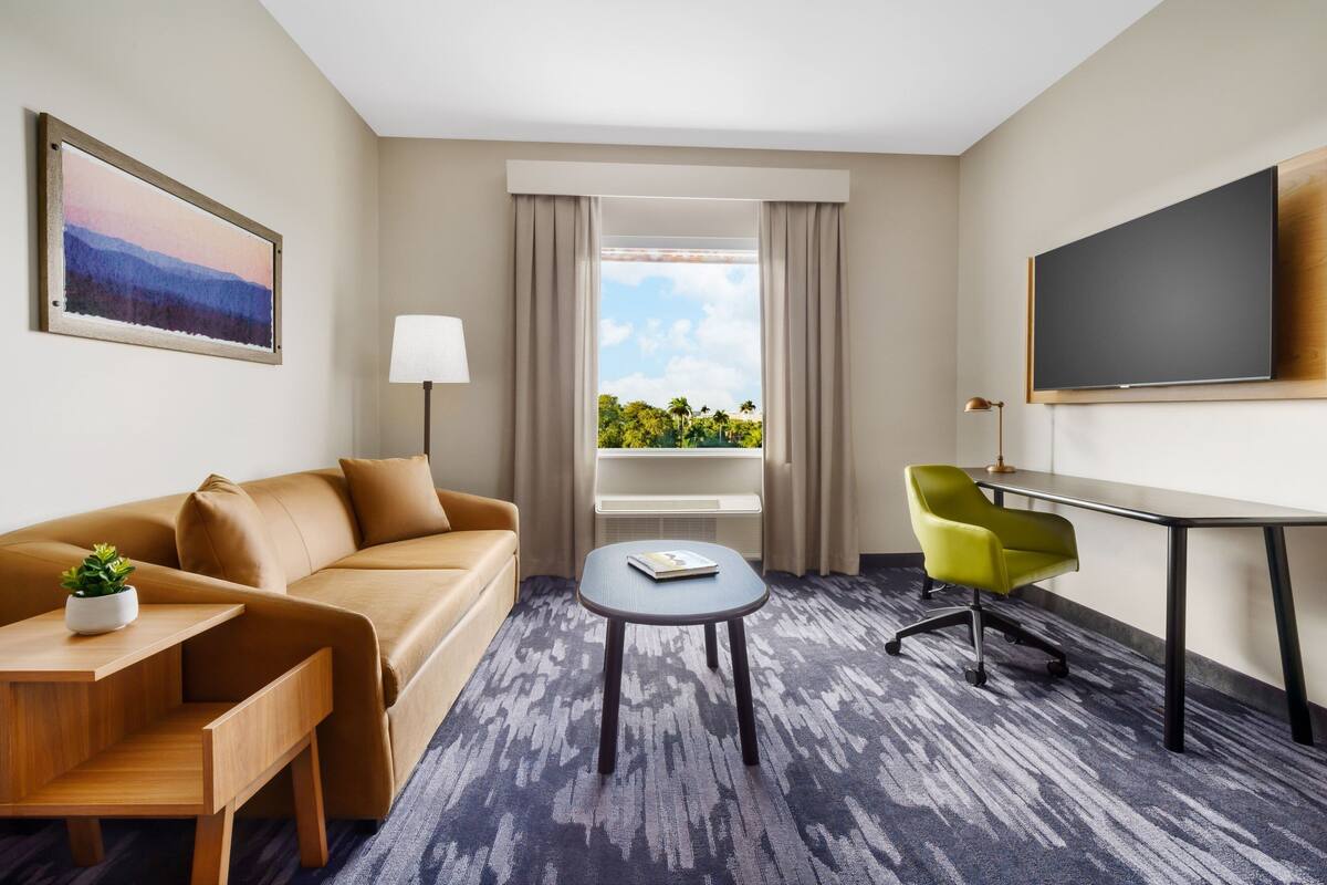 A hotel room with a sofa, a small table, a green chair, a TV, and a window with a view of greenery.