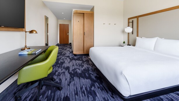 A hotel room with a bed, a desk, a green chair, and a television.