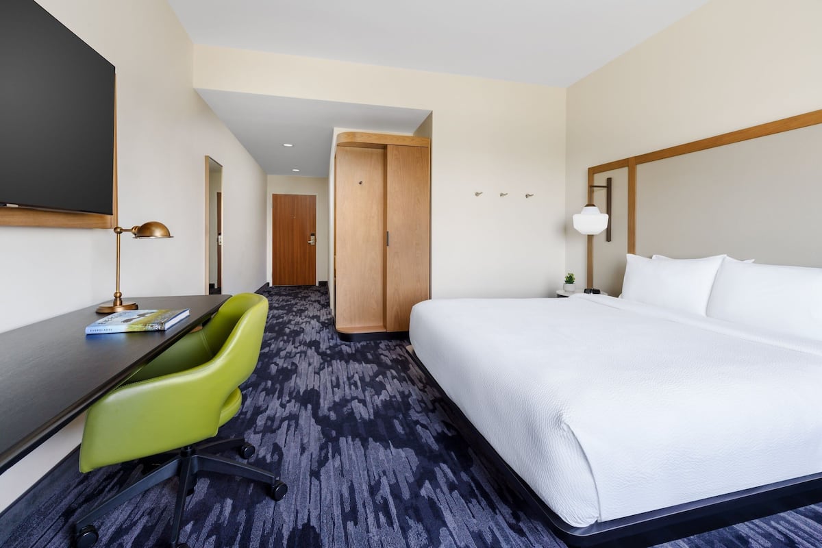 A hotel room with a bed, a desk, a green chair, and a television.
