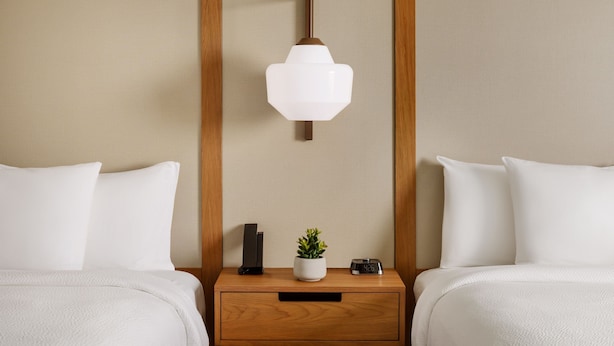 Habitación de hotel con dos camas, una mesita de noche con un reloj y una planta, y un aplique de pared.
