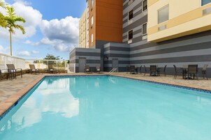 Una piscina con sillones de descanso y una palmera, rodeada por un edificio moderno de varios pisos y balcones.