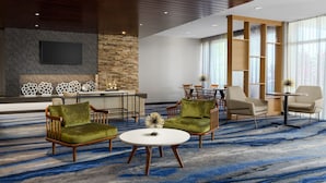 Un moderno vestíbulo de hotel con una pared de acento de piedra, un televisor de pantalla plana, una zona de estar con sillones tapizados en verde, una mesa de centro redonda y un gran ventanal con cortinas traslúcidas.