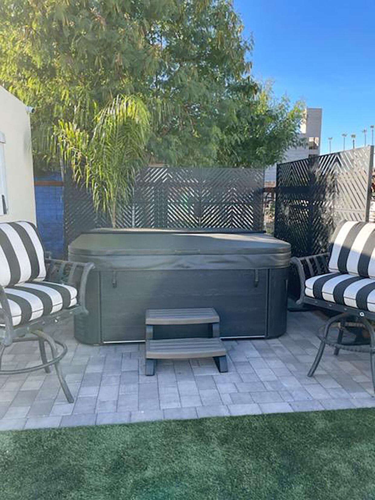 An outdoor patio with a hot tub, two striped chairs, and a small wooden stool.
