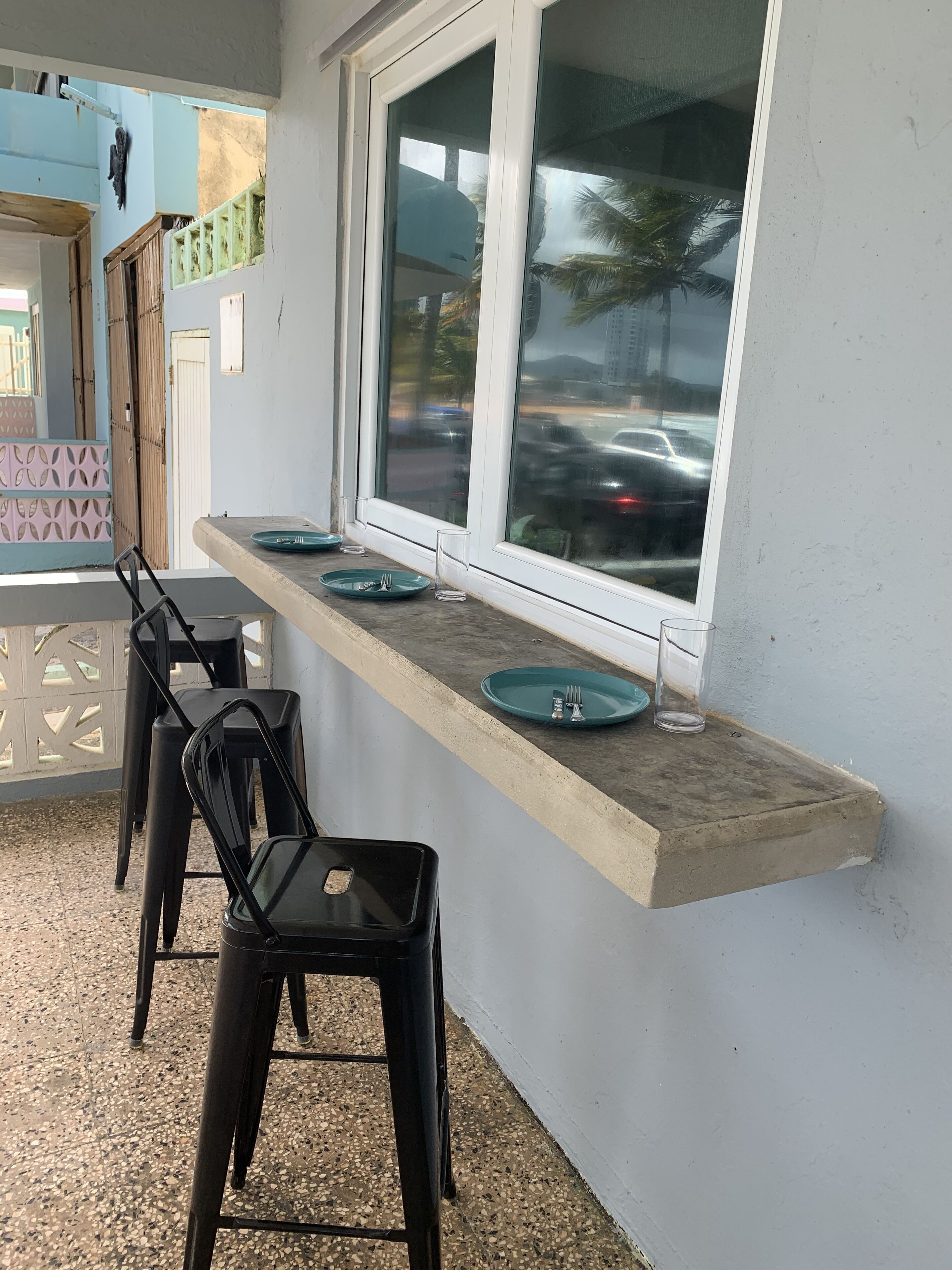 A small outdoor seating area with a concrete counter, black plastic chairs, and a window overlooking a street with cars.