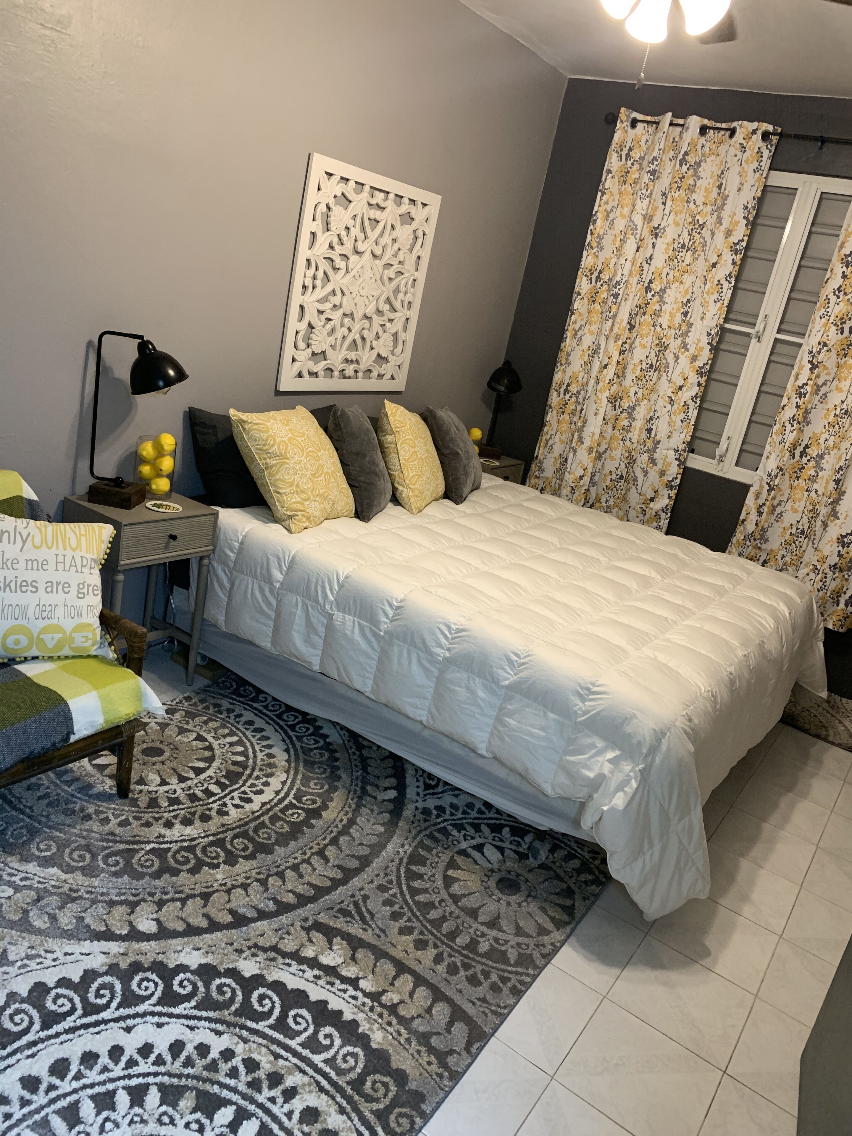 Une chambre à coucher avec un lit, une table de chevet, une lampe, une chaise, un tapis et une fenêtre avec des rideaux.