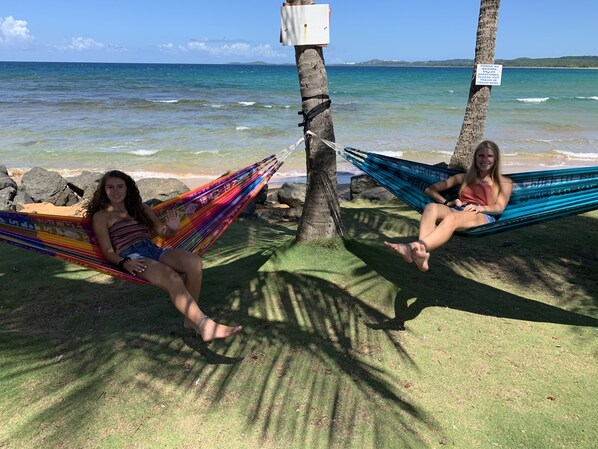 Deux personnes se détendent dans des hamacs suspendus à des palmiers, sur une plage bordée d’une eau claire et surplombant une terre lointaine.