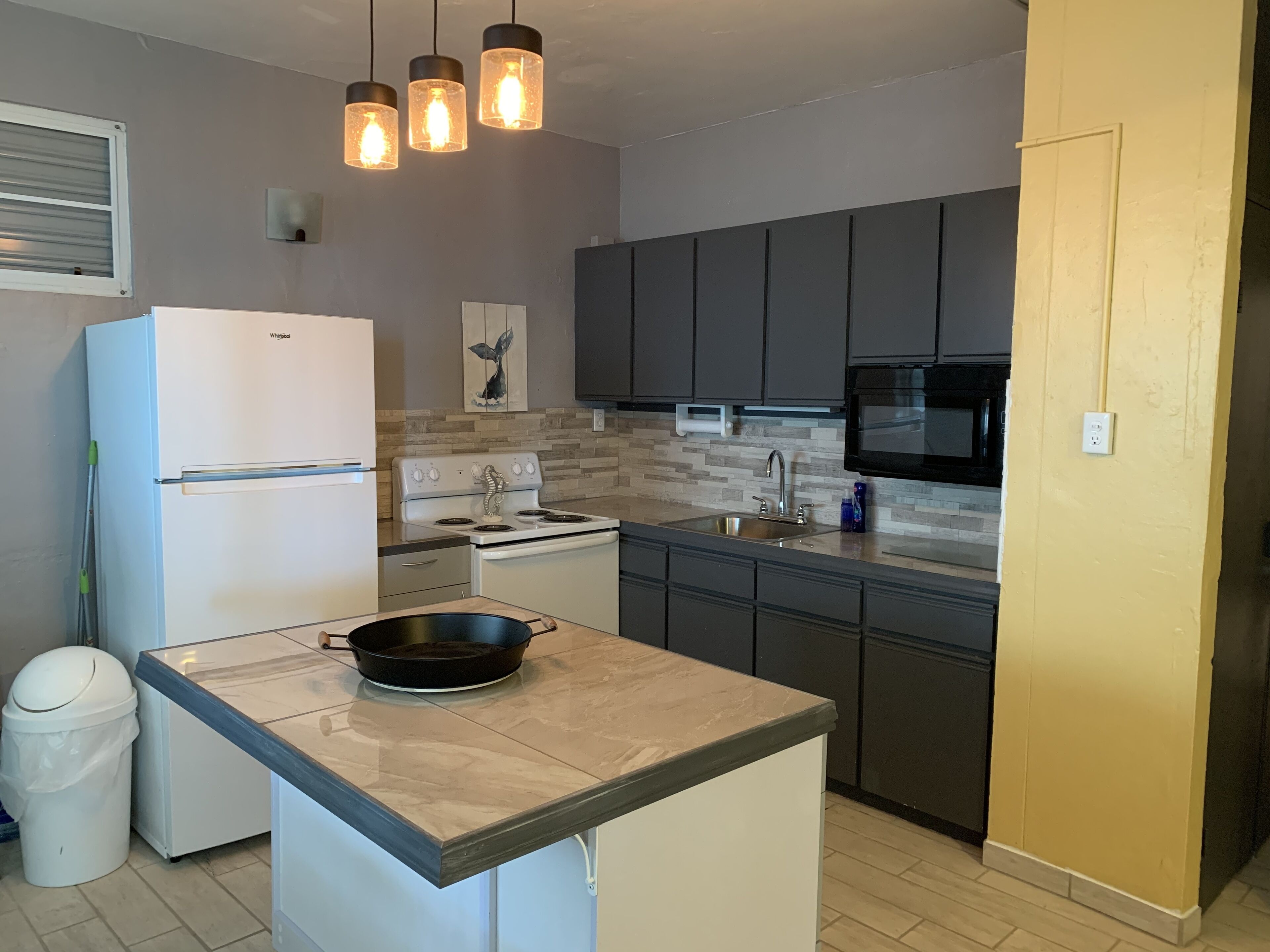 A modern kitchen with a white refrigerator, a stove, a sink, and a microwave. There are three pendant lights hanging above the counter.
