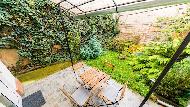 A covered patio with a wooden table and chairs, surrounded by lush greenery and a small garden.