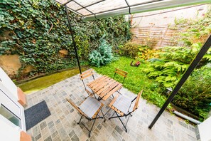Un porche cubierto con una mesa y sillas de madera, rodeado de abundante vegetación y un pequeño jardín.