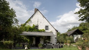 Una casa de pedra blanca amb teulada inclinada, un porxo cobert i un jardí amb plantes en testos.
