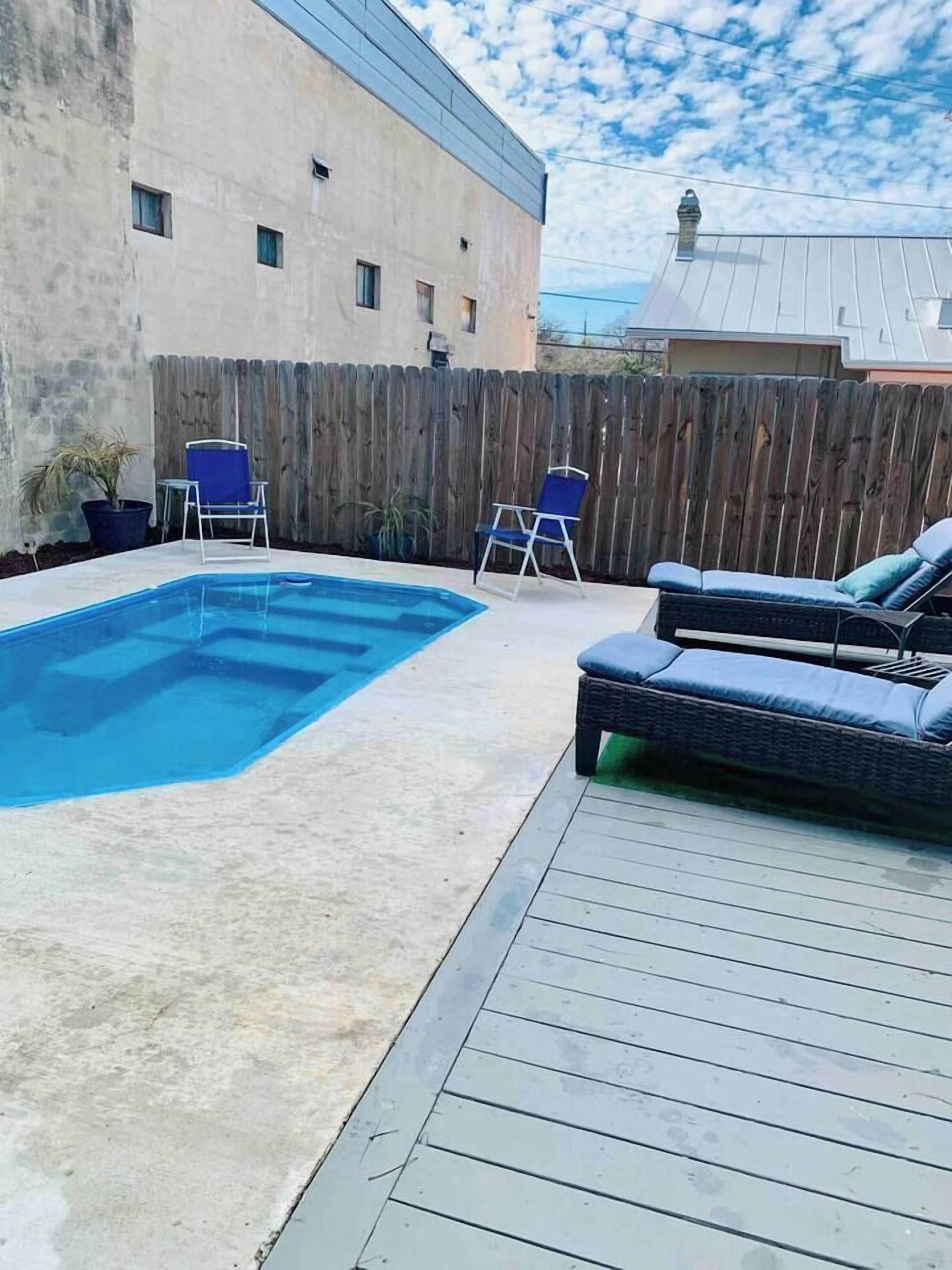A rectangular swimming pool with a wooden deck, two blue lounge chairs, and a wicker sofa.