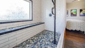 Une salle de bain aux murs recouverts de carreaux blancs, une fenêtre offrant une vue, une douche avec un tuyau noir et un lit avec une couverture blanche.