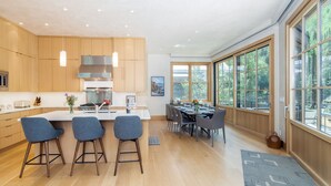 A modern kitchen with wooden cabinets, a central island with bar stools, and a dining area with a large window overlooking a forest. - THE Tributary by Exceptional Stays (Telluride)