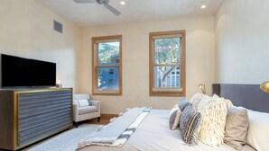 A bedroom with a bed, a TV on a wooden cabinet, two windows with a view of buildings, and a ceiling fan. - THE Tributary by Exceptional Stays (Telluride)
