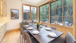 A modern dining area with a long wooden table set for six, large windows offering a view of a forested area, and a painting on the wall. - THE Tributary by Exceptional Stays (Telluride)