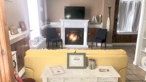 Una sala de estar con un sofá amarillo, una mesa blanca con un diploma enmarcado, una chimenea y un televisor encima.