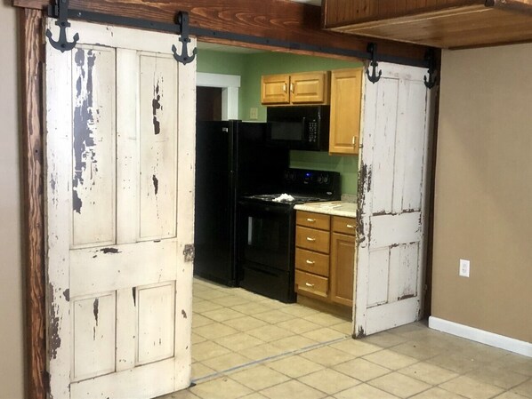 Una cocina con un refrigerador negro, microondas y estufa, armarios de madera y una puerta blanca desconchada.