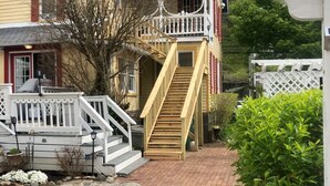 Una casa de dos pisos con una escalera de madera que lleva a un balcón, un camino de entrada empedrado y una cerca blanca de piquetes.