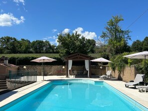 Piscina com espreguiçadeiras e guarda-sóis, cercada por um muro de tijolos e árvores.