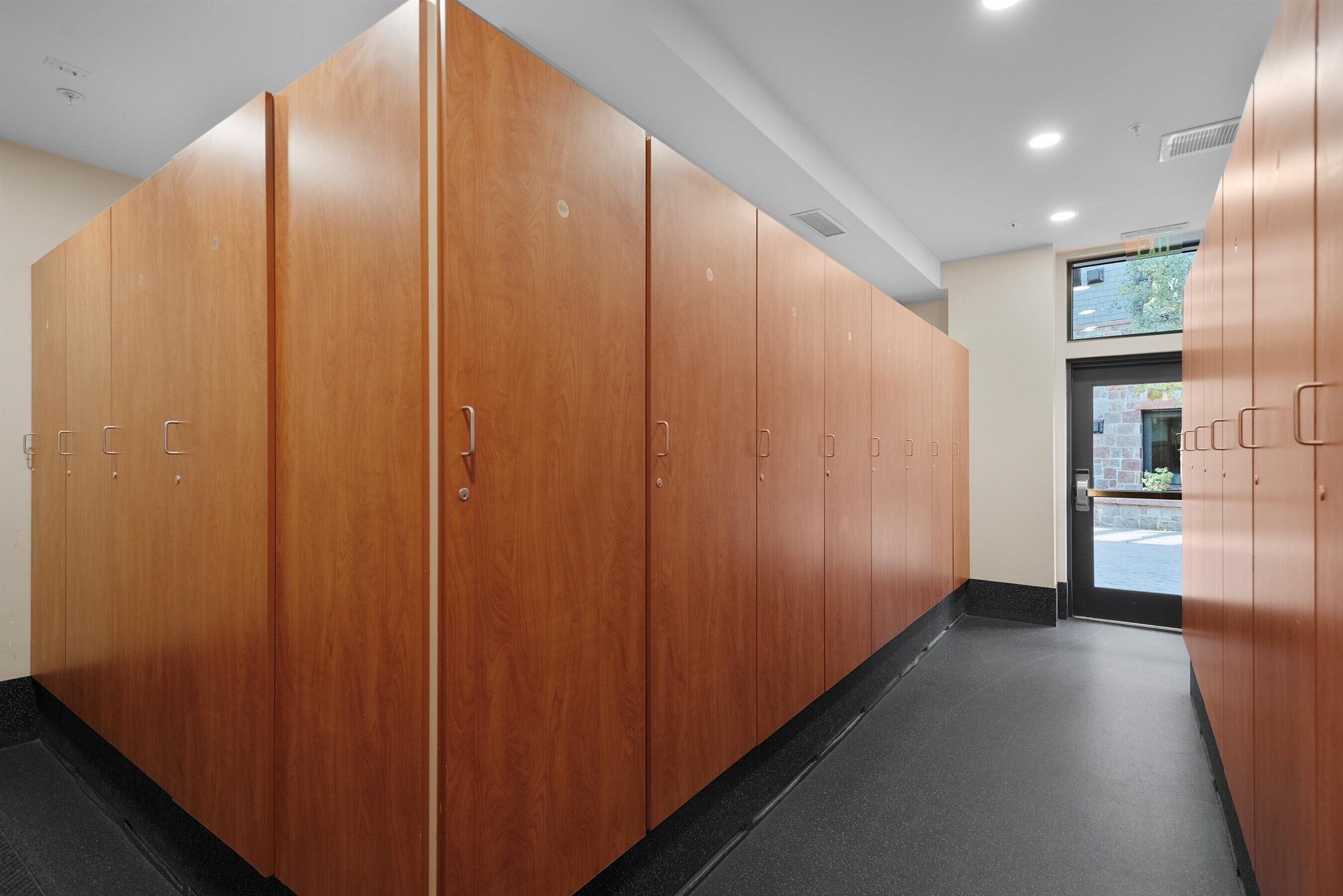 A row of wooden lockers with metal handles, a black floor, and a door leading outside.