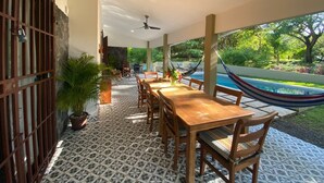 A covered patio with a dining table and chairs, a hammock, and a view of a swimming pool and greenery.