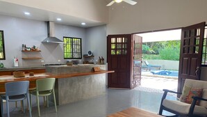 A modern kitchen with a central island, dining area with chairs, and a view of a swimming pool through an open door.