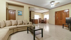 Un salon avec un canapé beige, une table basse et un téléviseur posé sur un meuble. On aperçoit à l’arrière-plan un coin repas avec une table et des chaises.