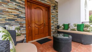 Une porte en bois, un mur décoré d’une mosaïque de pierres, des meubles en osier et une table en verre.