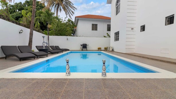 Une piscine avec des chaises longues, un barbecue et un bâtiment blanc avec un toit rouge en arrière-plan.